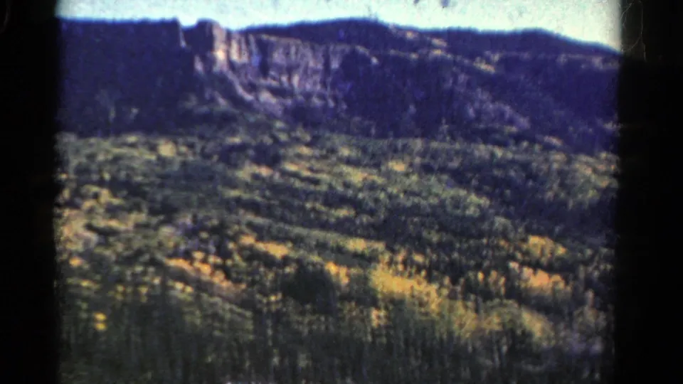 1967 Colorado landscape frame showing a broad evergreen valley, sunlit rocky cliff, dark ridge line, and pale sky on 8mm film.