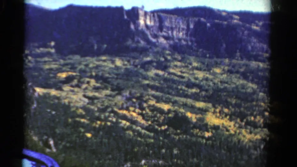 1967 Colorado 8mm frame showing a forested mountain valley beneath a pale cliff band and distant ridge.