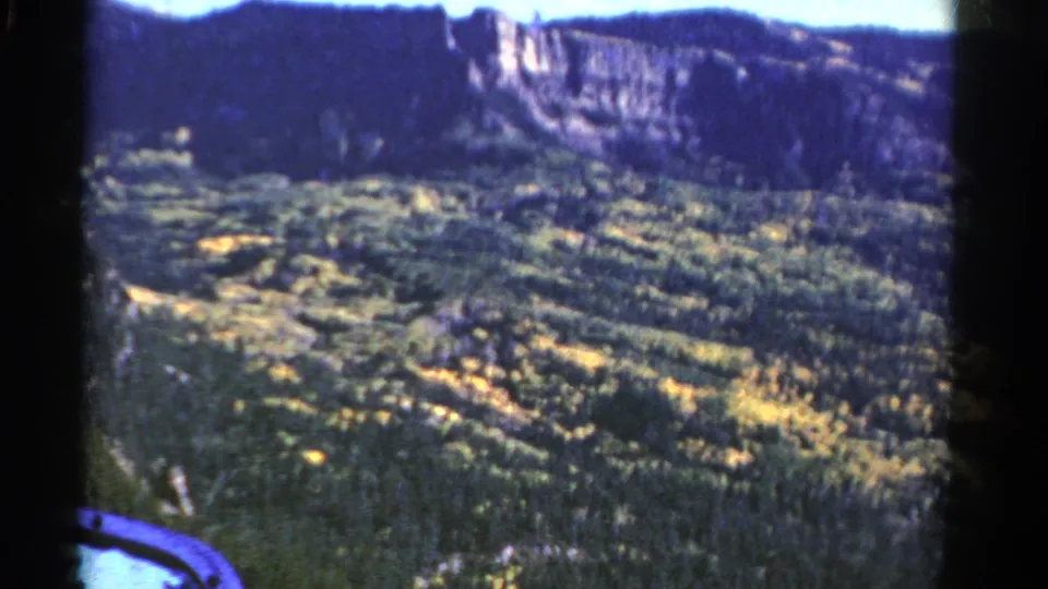 1967 Colorado home-movie view across a wooded valley toward a rugged cliff wall and dark ridge line.