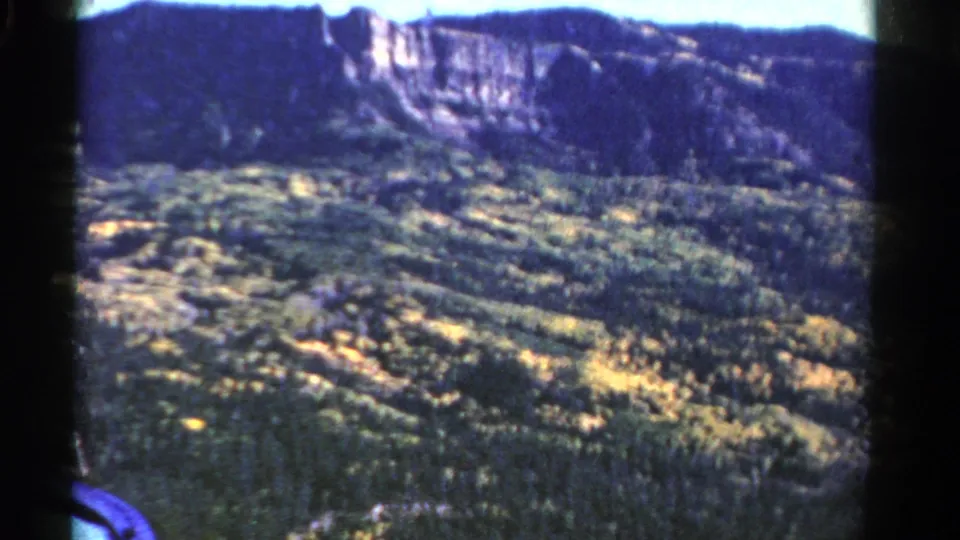 1967 Colorado 8mm frame with soft focus on forested slopes, autumn-colored patches, and a distant rock cliff.