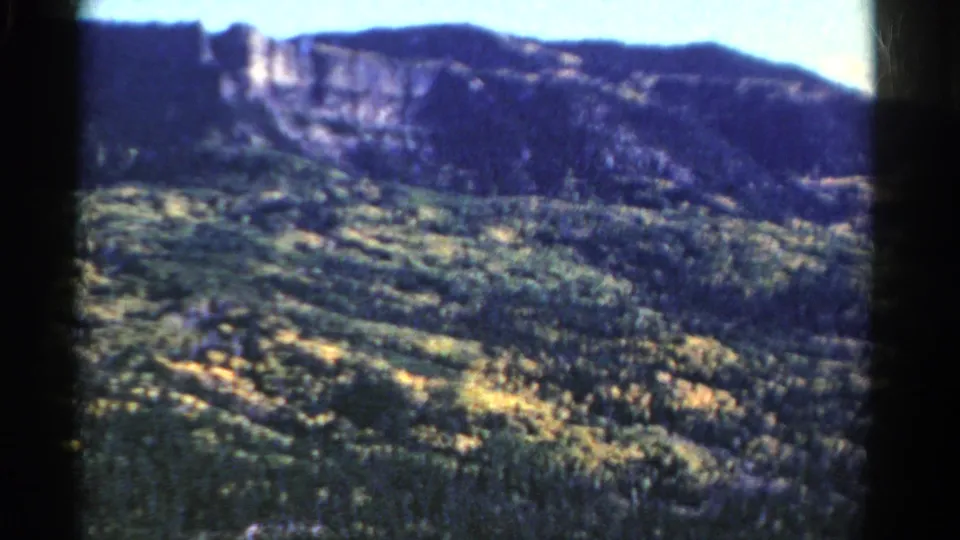 1967 Colorado home-movie frame looking over trees and yellow-green ground cover toward a cliff face and ridge.