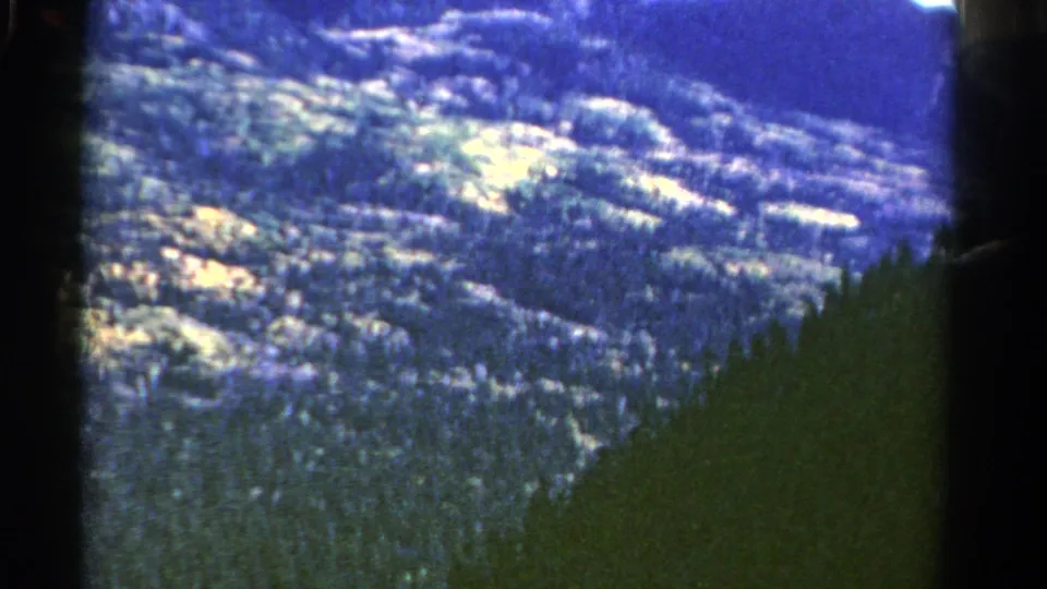 1967 Colorado 8mm aerial view with a dark foreground hillside, forested ridges, and bright patches of vegetation.