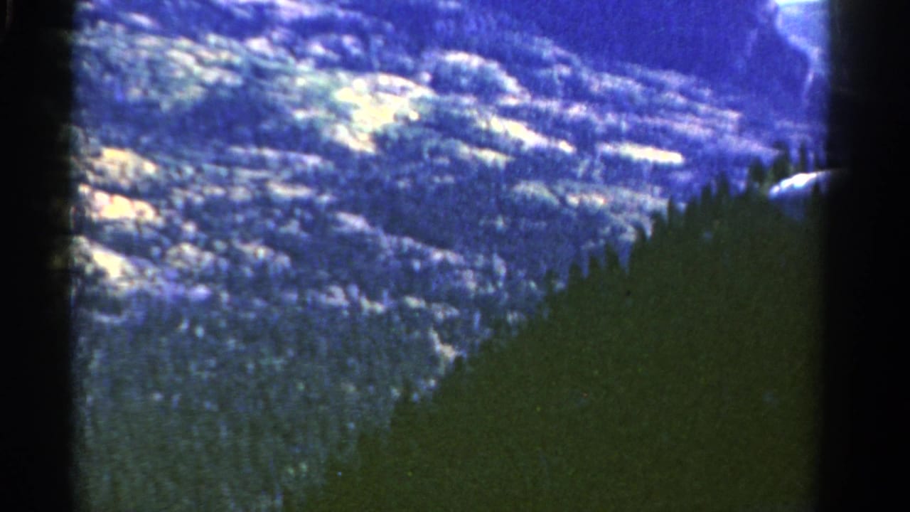 Distant snowy mountain slopes above dark evergreen forest with film frame edges visible.