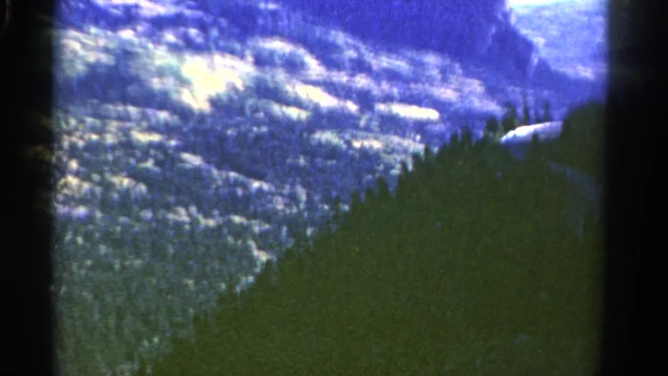 Colorado mountain scenery on 8mm film, showing a forested ridge below pale snow patches and distant rock forms.