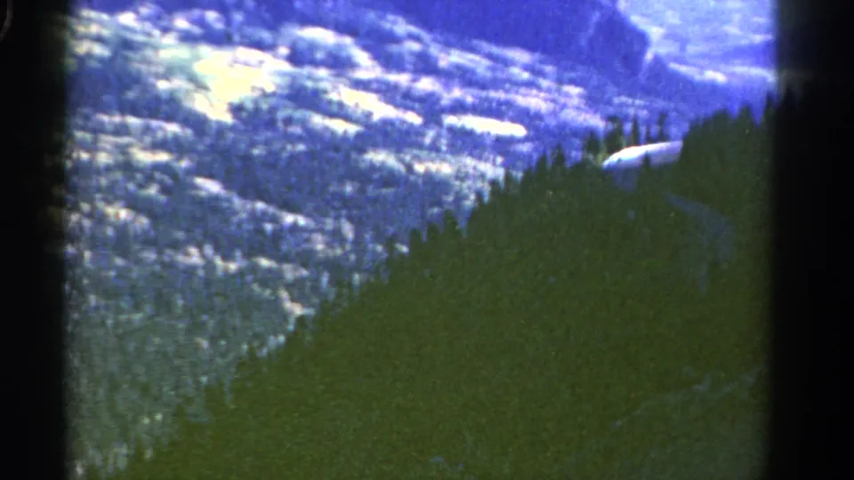 1967 Colorado home-movie frame of a mountain valley, evergreen slopes, and a faint road curving through the lower terrain.