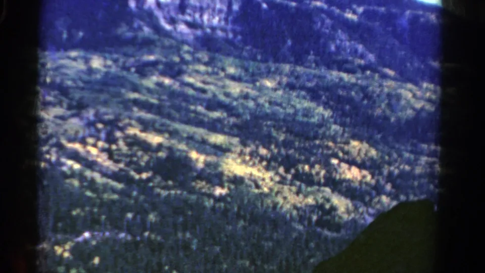Vintage 8mm Colorado aerial frame showing rolling forested terrain, pale sunlit clearings, and cliff rock in the distance.
