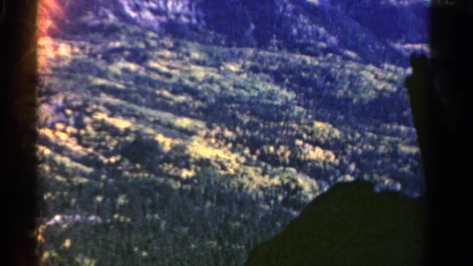 1967 Colorado 8mm frame of wooded mountain terrain beneath rocky high ground, with dark film border visible along the side.