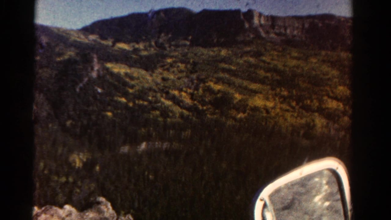 Mountain hillside and green valley seen past a car side mirror.