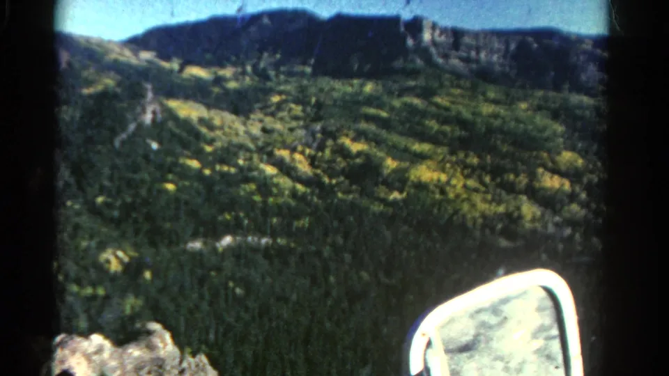 Colorado hillside and forested slope filmed from a moving vehicle in 1967, with rocks and a bright side mirror in view.