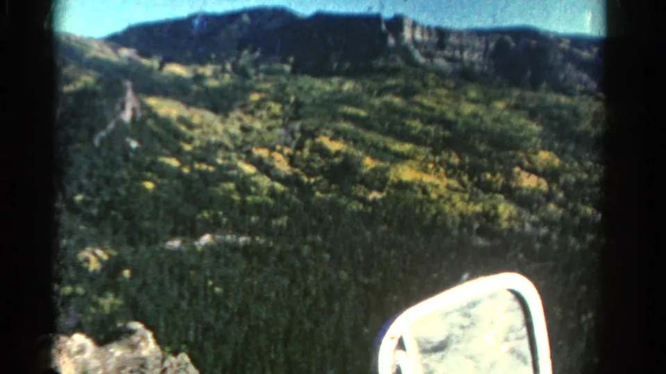 1967 Colorado car-window view of a sunlit hillside below a dark ridge, with 8mm softness and side mirror visible.