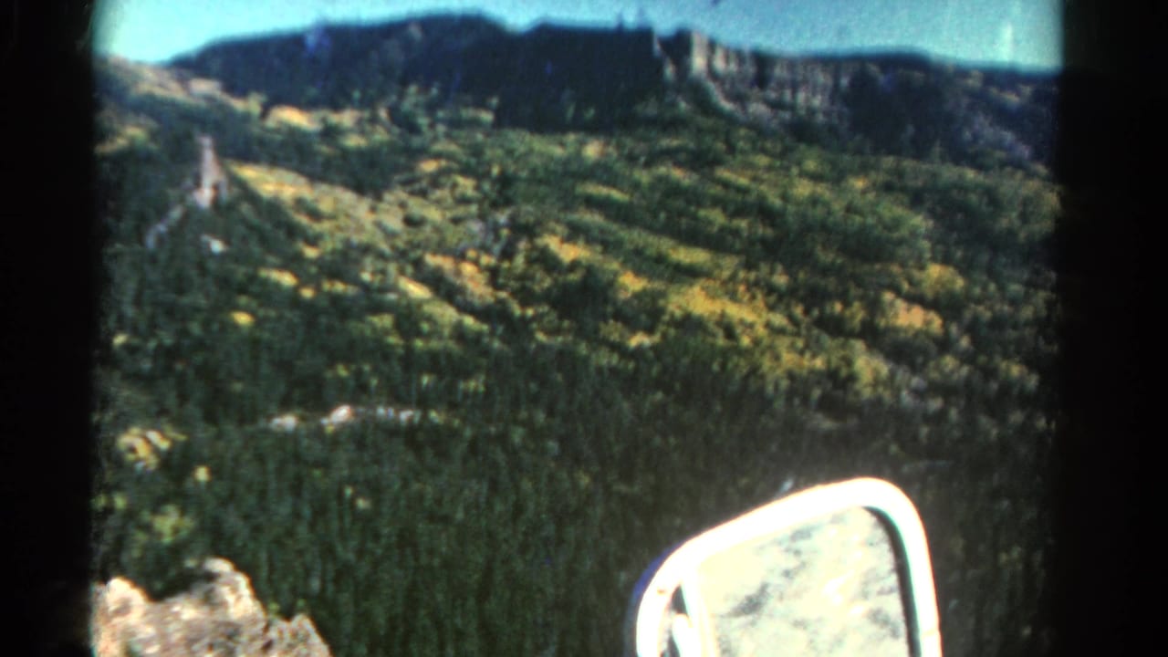 1967 Colorado 8mm frame showing a green mountain valley, yellow tree patches, distant rocky ridge, and car side mirror at the lower right.