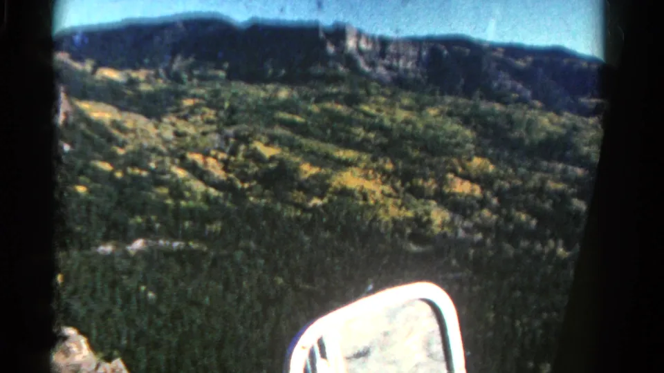 1967 Colorado travel footage frame with a bright car side mirror below a green valley, yellow tree patches, and a distant rocky ridge.