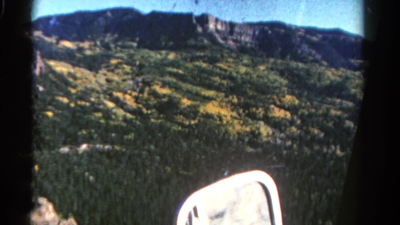1967 Colorado 8mm frame showing a forested mountain slope, blue sky, dark ridge line, and a white car side mirror from a passenger viewpoint.