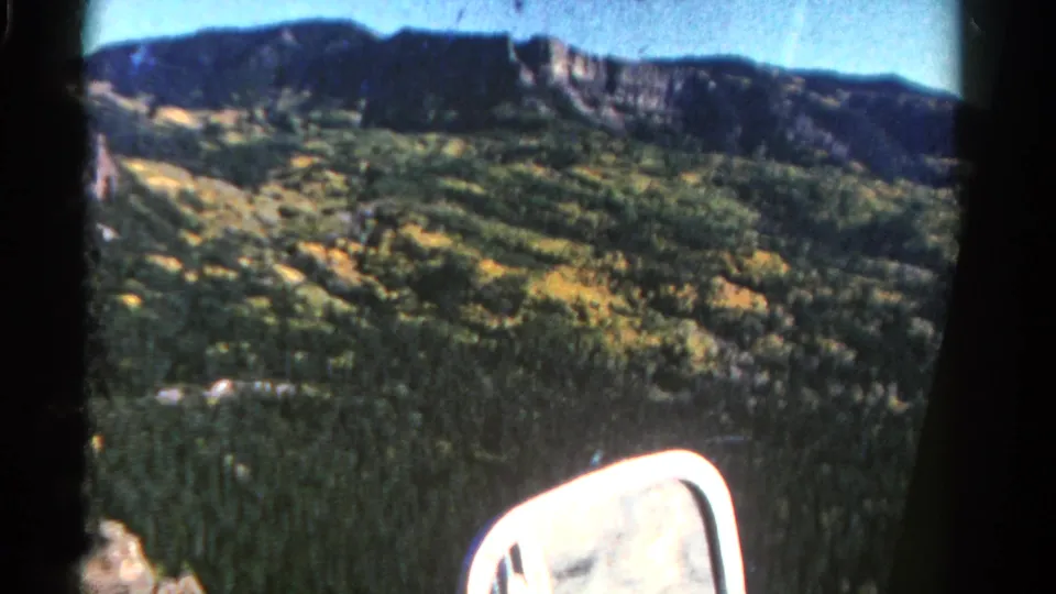 1967 Colorado 8mm frame showing a forested mountain slope, blue sky, dark ridge line, and a white car side mirror from a passenger viewpoint.