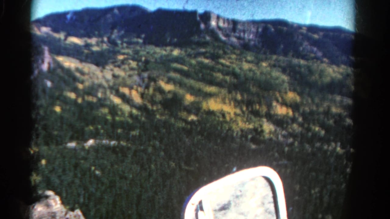 1967 Colorado road-view frame with evergreen-covered slopes, scattered yellow foliage, a dark ridgeline, and the car side mirror at lower right.