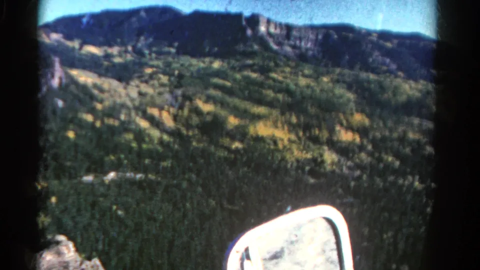 1967 Colorado road-view frame with evergreen-covered slopes, scattered yellow foliage, a dark ridgeline, and the car side mirror at lower right.
