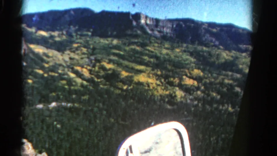 1967 Colorado 8mm frame with soft motion blur over forested hills, bright foliage patches, distant ridge, and a white car side mirror.