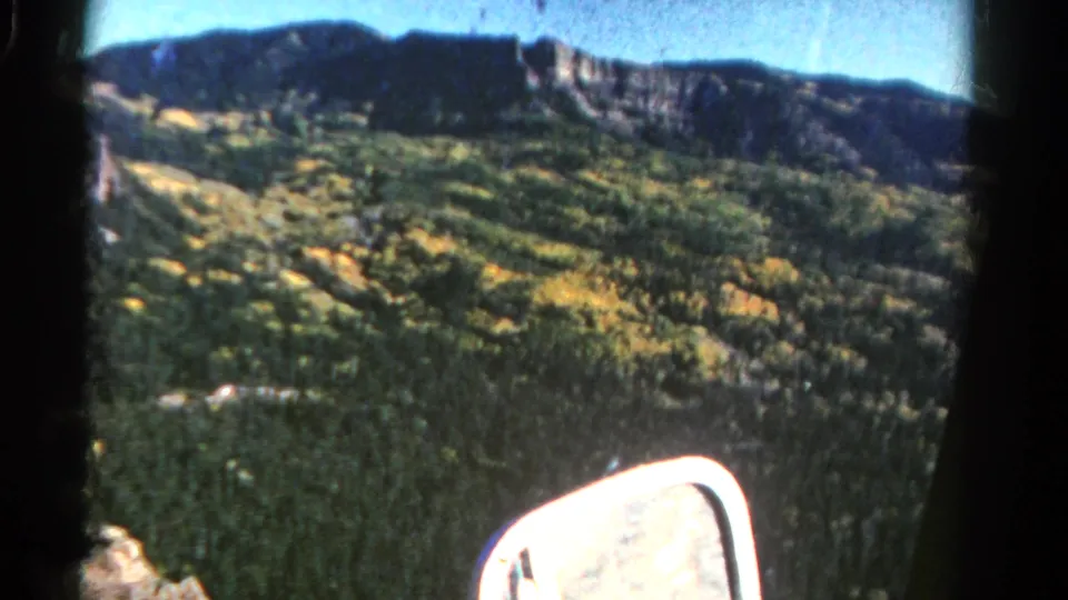 1967 Colorado home-movie frame showing dense mountain forest, yellow foliage patches, a dark ridge, blue sky, and the vehicle side mirror.
