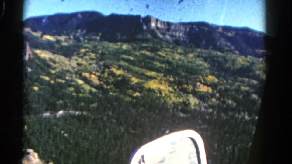1967 Colorado 8mm frame showing a tree-covered mountain ridge and autumn-toned slopes through a moving vehicle window.