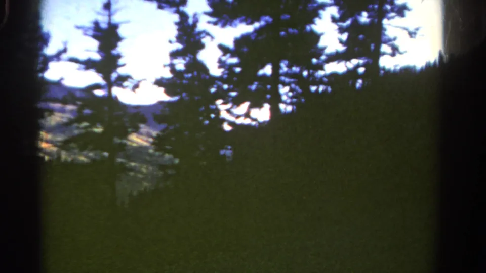 1967 Colorado 8mm frame showing evergreen silhouettes, shadowed hillside, pale sky, and distant mountain shapes.