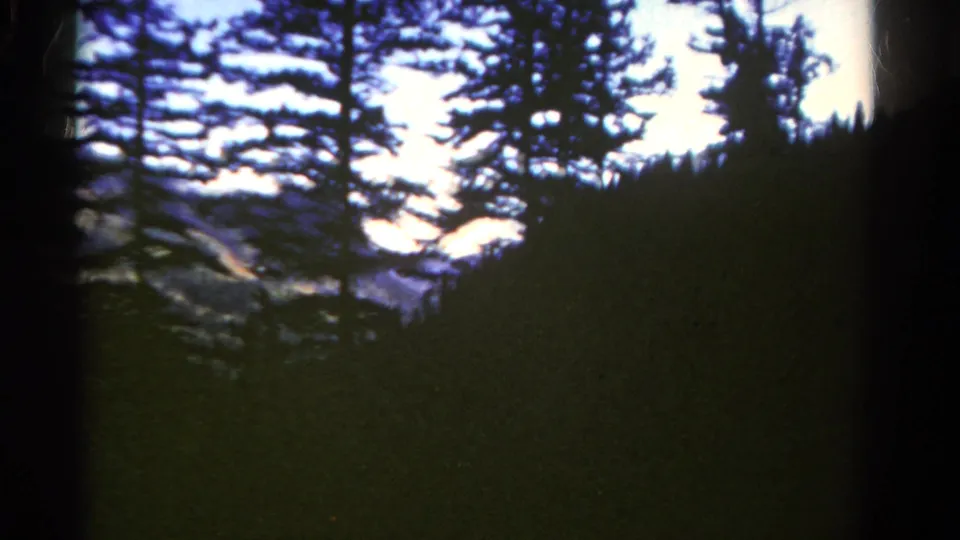 1967 Colorado frame showing a dark forested hillside, silhouetted trees, pale sky, and distant mountain forms.