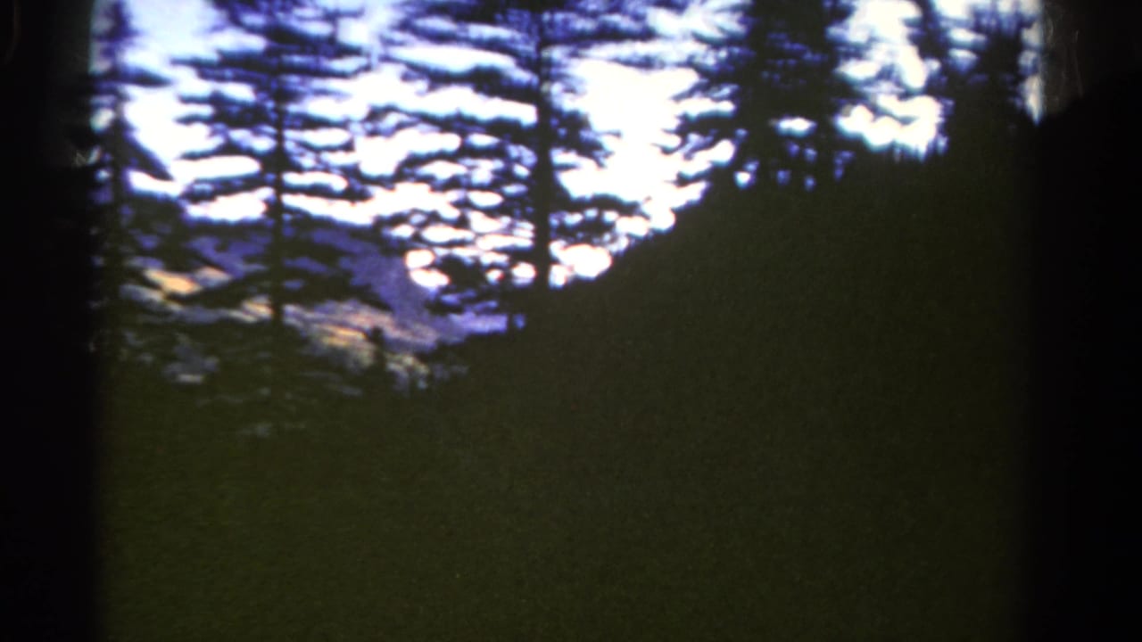 1967 Colorado home-movie frame with pine silhouettes, wooded mountain slopes, and a pale sky.