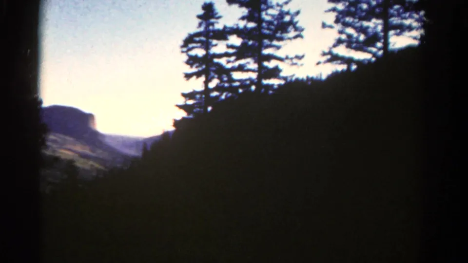 1967 Colorado travel frame showing a dark wooded slope, pine silhouettes, and a distant mesa-like ridge.