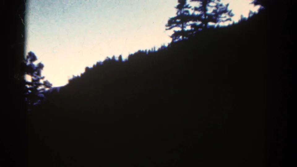 Colorado 8mm frame with a shadowed mountain slope, scattered pine silhouettes, and pale open sky.
