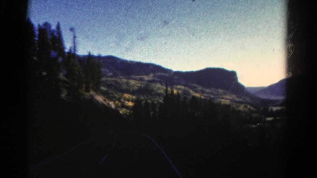 1967 Colorado 8mm frame showing a dark curving road, pine forest, distant mountain ridges, and a brightening morning sky.