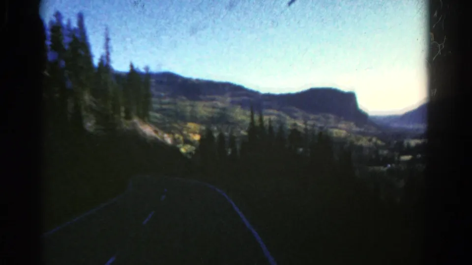 1967 Colorado 8mm frame showing a shadowed road foreground, pine trees, sunlit slopes, and a pale blue sunrise sky.