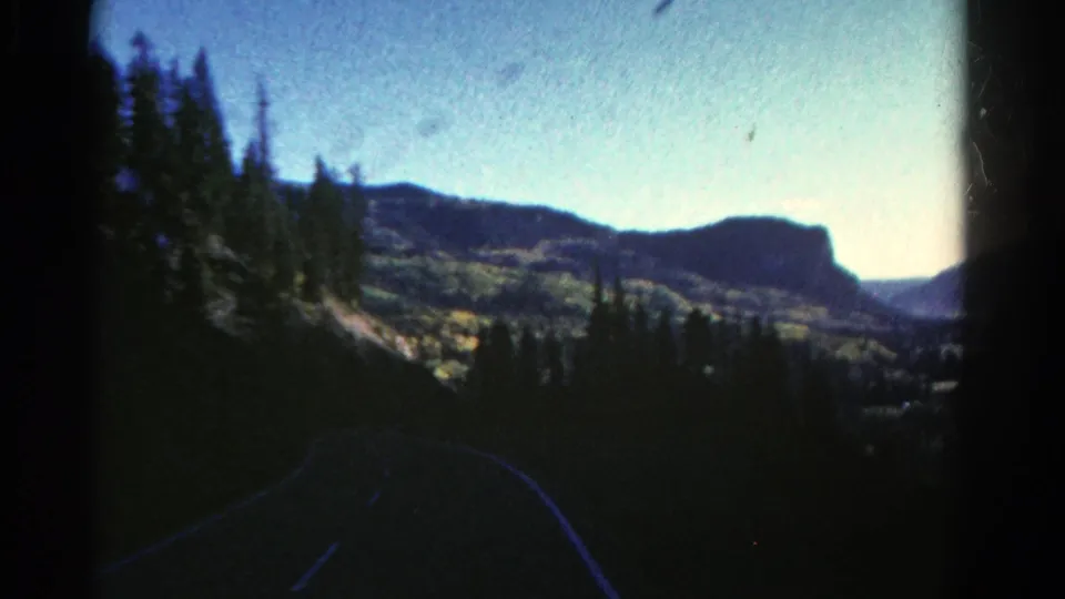 1967 Colorado home-movie frame with a curved mountain road, dark evergreen trees, distant ridges, and bright sunrise exposure.