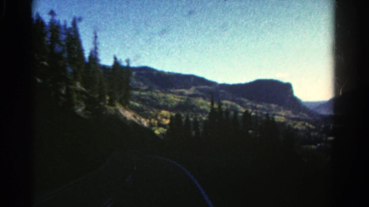 Mountain road view with pine trees, dark foreground, distant mesa, and pale blue sky.