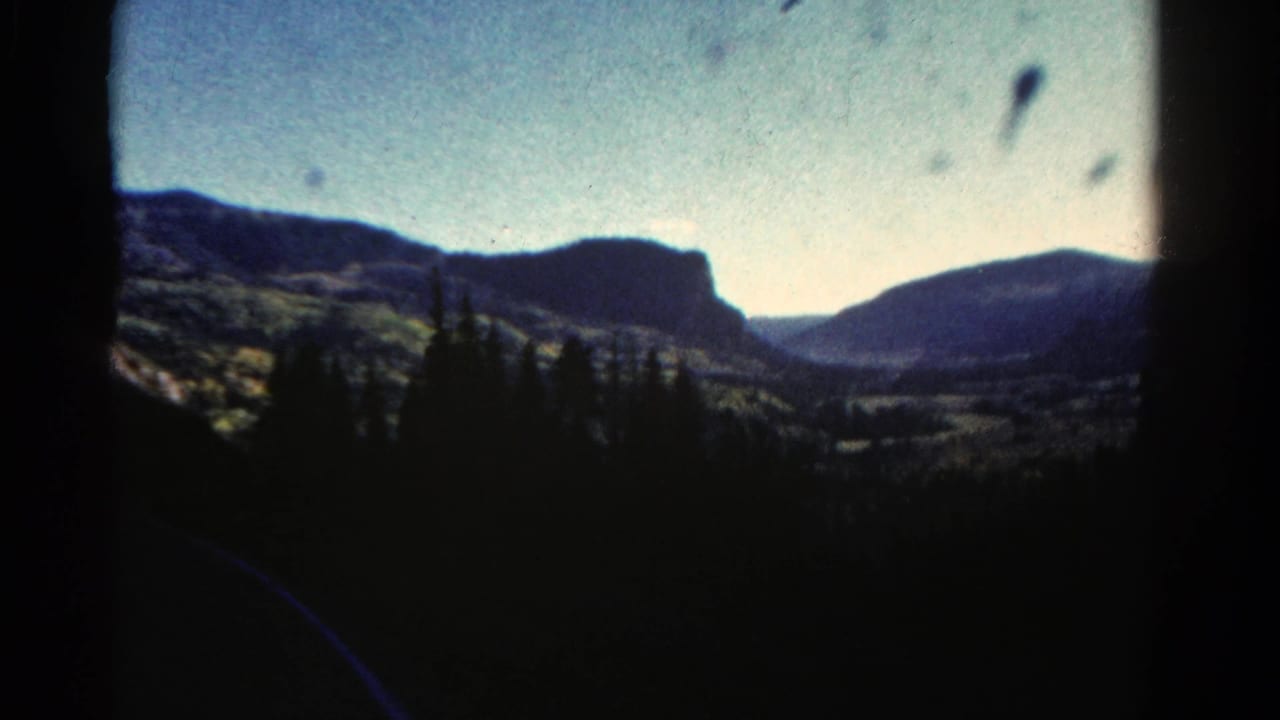 1967 Colorado 8mm frame of dark pine silhouettes in front of distant mountain ridges and a valley under pale sky.