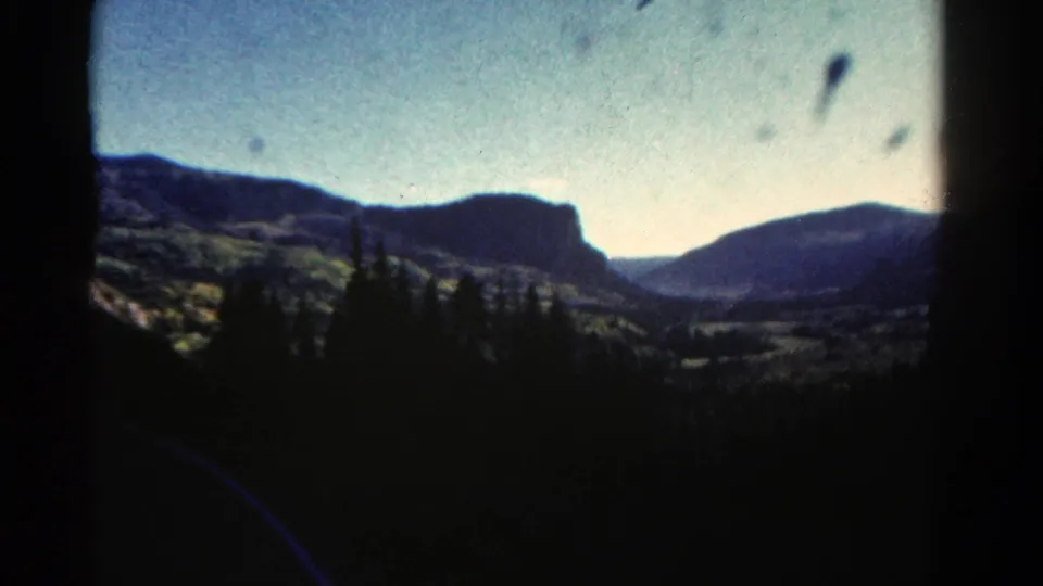 1967 Colorado 8mm frame of dark pine silhouettes in front of distant mountain ridges and a valley under pale sky.