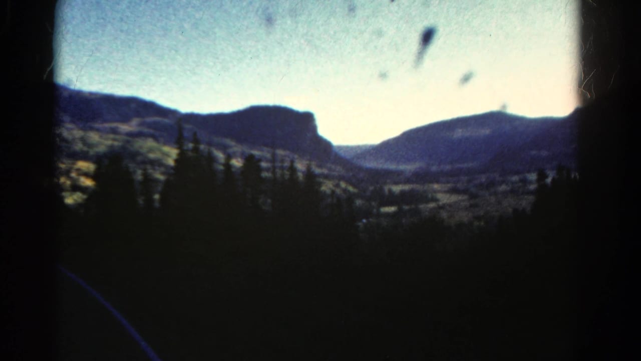 1967 Colorado home-movie frame showing a soft-focus mountain valley beyond dark evergreen trees and film edge shadows.