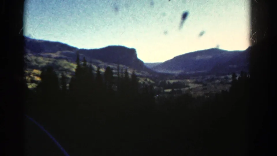1967 Colorado home-movie frame showing a soft-focus mountain valley beyond dark evergreen trees and film edge shadows.