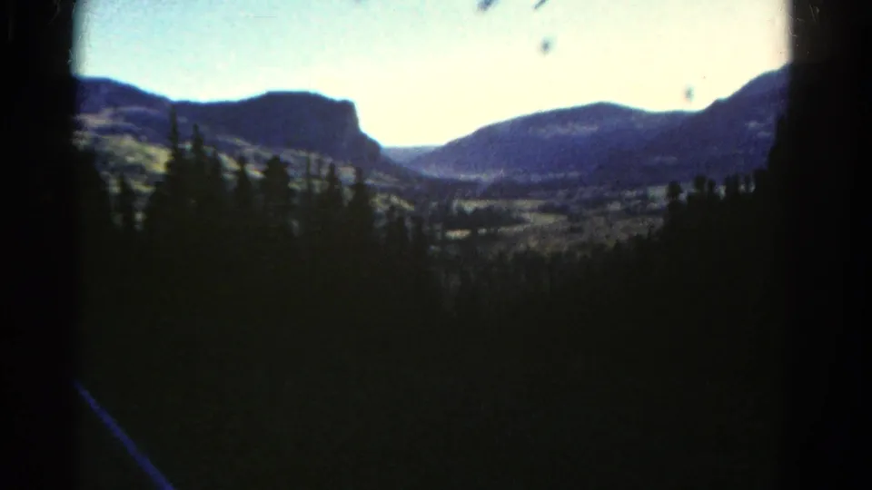 1967 Colorado 8mm frame with blurred pine foreground, distant ridges, pale sky, and visible home-movie film artifacts.