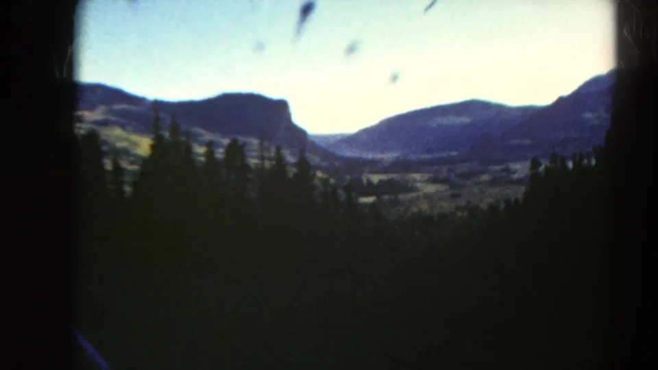 1967 Colorado home-movie frame of a pine-covered foreground, distant valley, and mountain ridges beneath a bright sky.