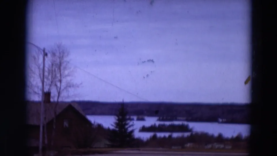 1967 8mm Detroit-area home-movie frame showing a lakeside house, bare trees, an evergreen, power line, water, and distant islands.