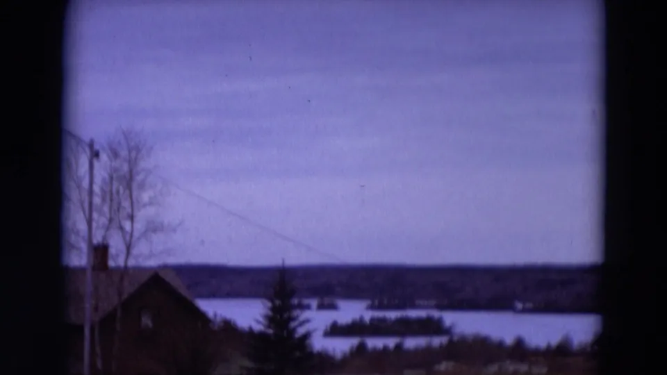 1967 8mm Detroit-area frame with a house partly low in frame, bare trees, evergreen, lake water, and distant islands.