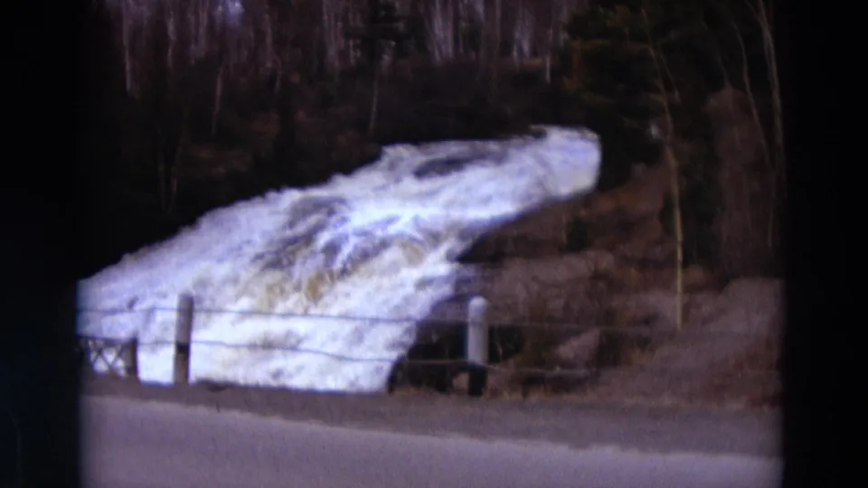 Detroit-area 1967 home-movie frame showing bright rushing meltwater or river, snowbanks, fence posts, road edge, and leafless woods.