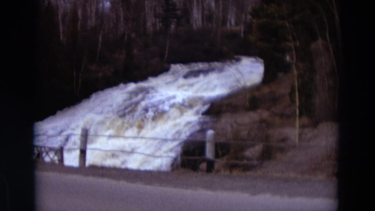 Turbulent white water spills beside a road, with posts, wire fencing, and bare trees in the background.