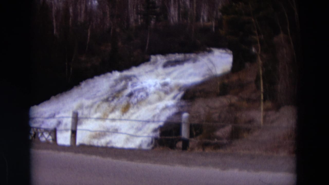 Rushing water continues past roadside posts and a low wire fence, framed by dark film edges.