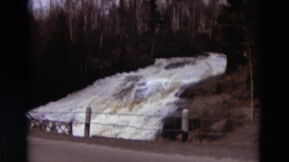 Soft 1967 Detroit-area home-movie frame showing overflow water, roadside pavement, fence posts, and bare trees.