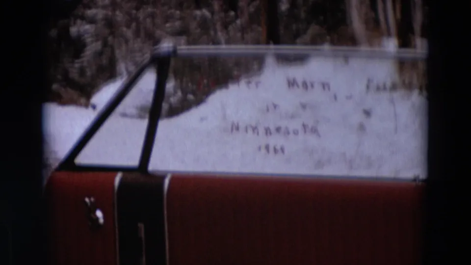 Red car stopped near the roadside with handwritten windshield text reading Detroit Michigan 1967, seen in soft 8mm focus.