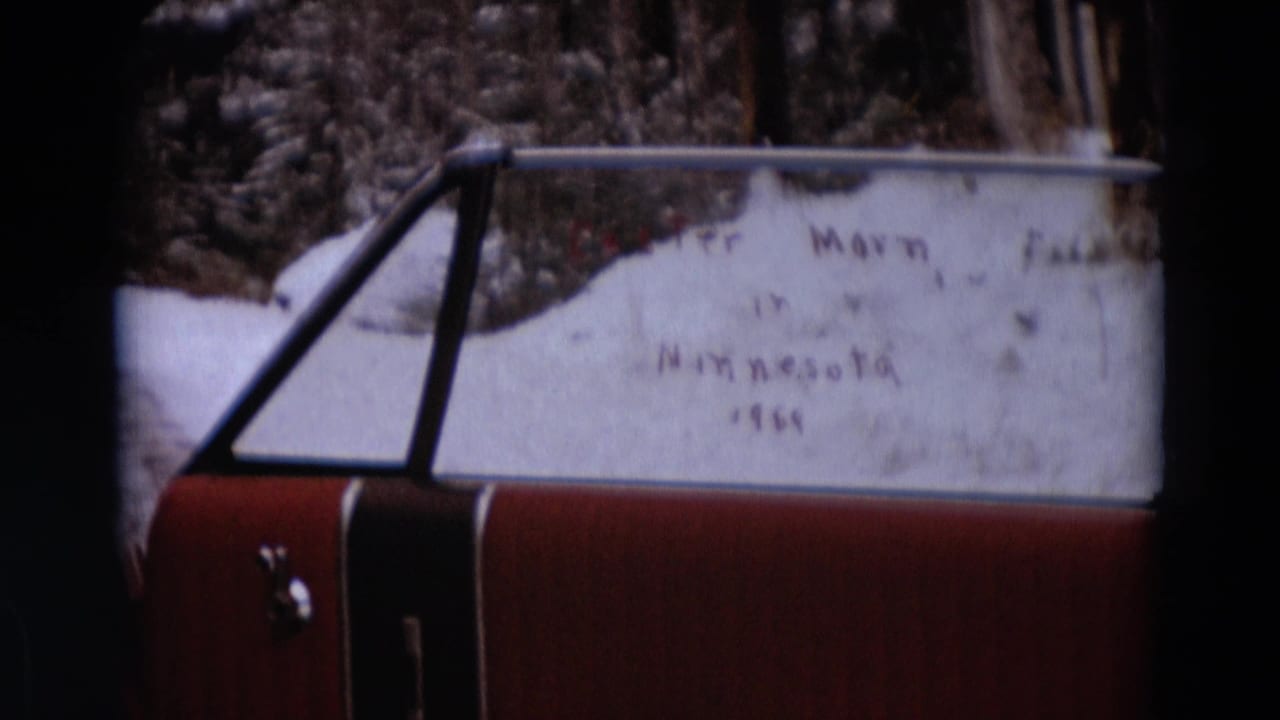 1960s 8mm frame of a red car in snow, showing handwriting on a snow-covered window and bare trees in the background.