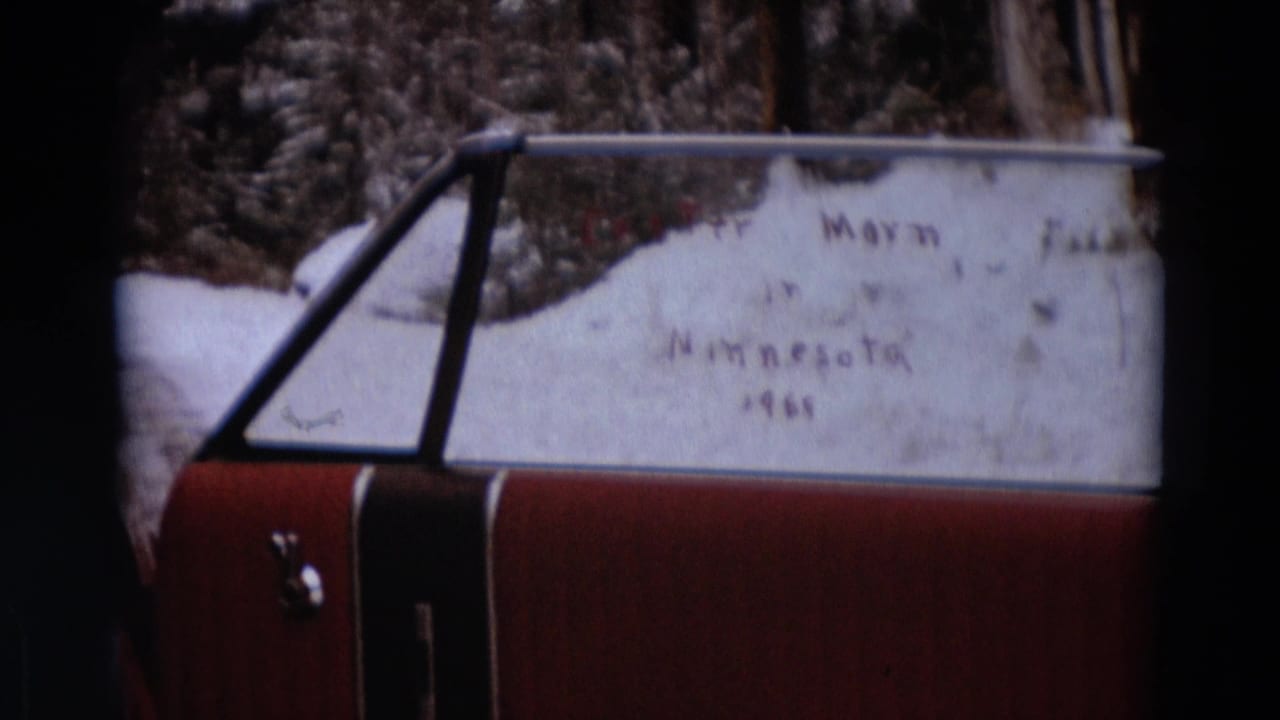 Detroit-area home-movie frame showing a red car window with snow and faint handwritten wording visible against a wooded winter background.