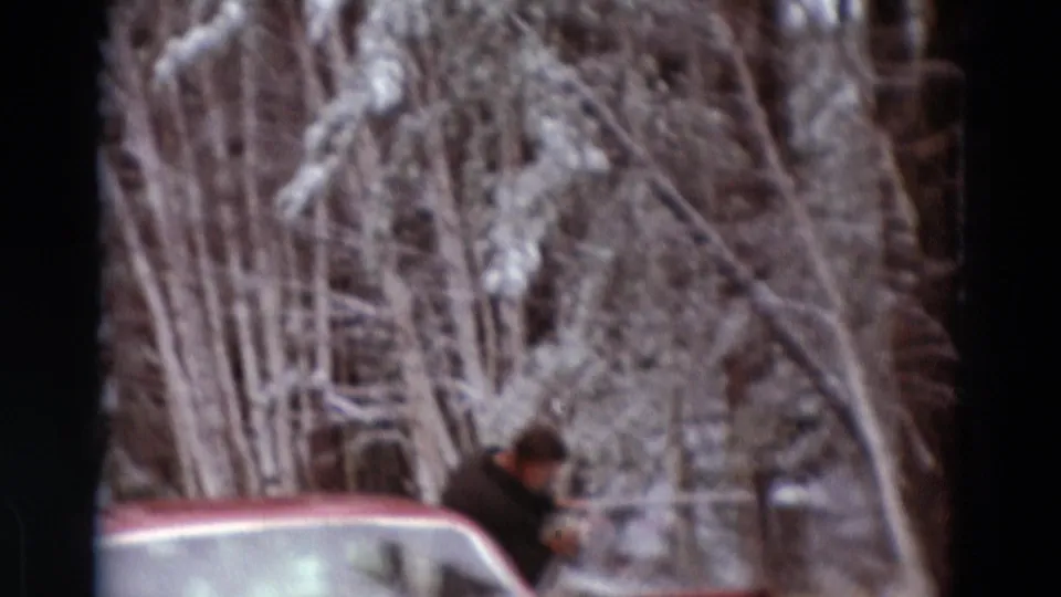 A person in dark clothing appears beside the red car, with snowy trees behind and the soft grain of 8mm film visible.