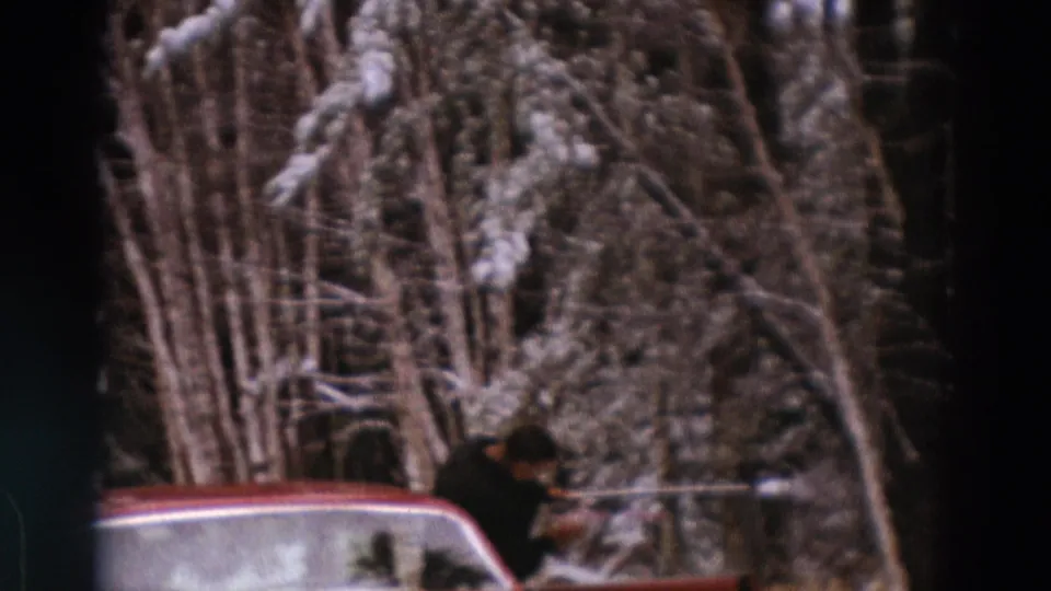1960s home-movie frame of a person using a long-handled tool by a red car, with snow-covered trees behind.