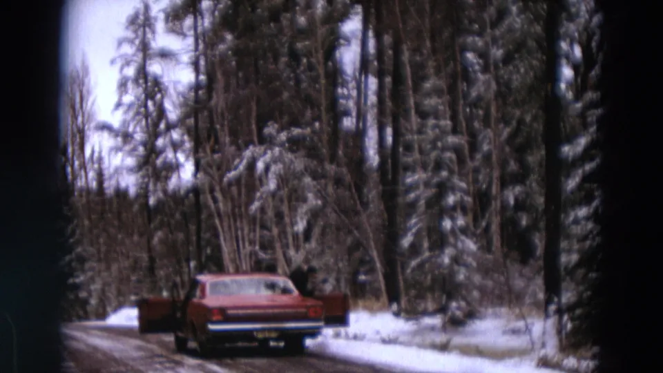 A person in dark winter clothing stands at a red sedan on a snow-lined road in the Detroit, Michigan clip metadata context.